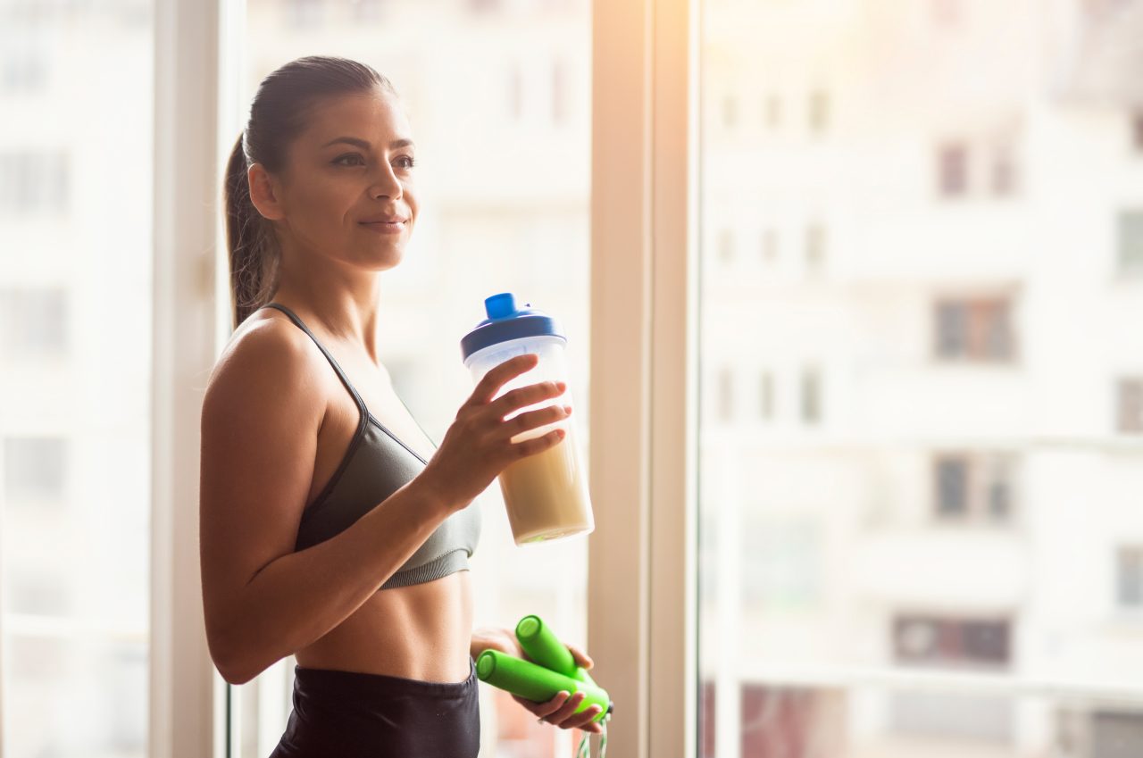 Bodybuilder girl relax after exhausting training, Young athlete drinking sports drink after workout, beautiful woman resting after exercising training, young sports model holding protein shaker and skipping rope around the window.